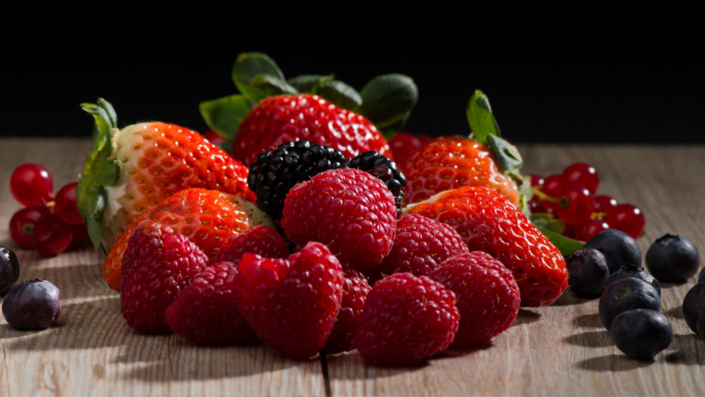 Fruits rouges posés sur une table en bois foncé avec un fond noir. Fraises, framboises, groseilles et cassis