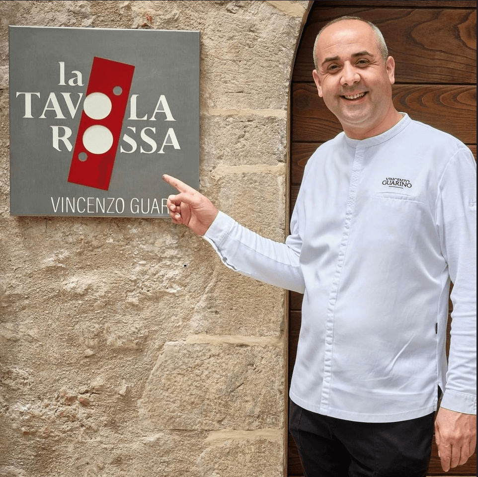 Immagine che mostra lo chef Vincenzo Guarino mentre indica la targa del suo ristorante La Tavola Rossa presso la Teritoria Castello di Postignano.