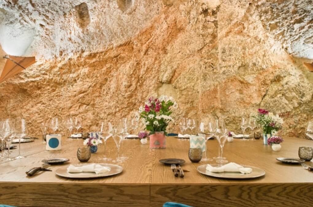 Vue d'une des tables du restaurant La Tavola Rossa au sein de Castello di Postignano. Large table en bois entourée de murs en pierre et habillée de façon élégante.